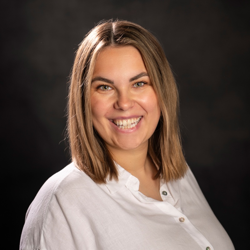 Sabrina Geibel (BWL - Bachelor of Science) mit schulterlangem braunem Haar lächelt, trägt eine weiße Bluse und steht vor einem dunklen Hintergrund.