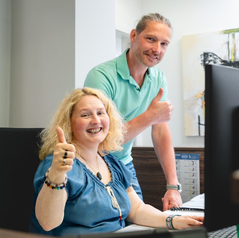 Bilanzbuchhalter Steuerfachwirt Eine Mitarbeiterin und ein Mitarbeiter der Steuerkanzlei Möller im Büro zeigen Daumen hoch und lächeln vor einem Computerbildschirm.
