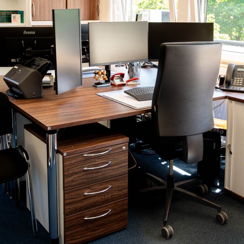 Ein modernes Büro mit mehreren Monitoren, einem Schreibtischstuhl, Telefon und Büromaterialien auf einem Holztisch in hell beleuchtetem Raum.