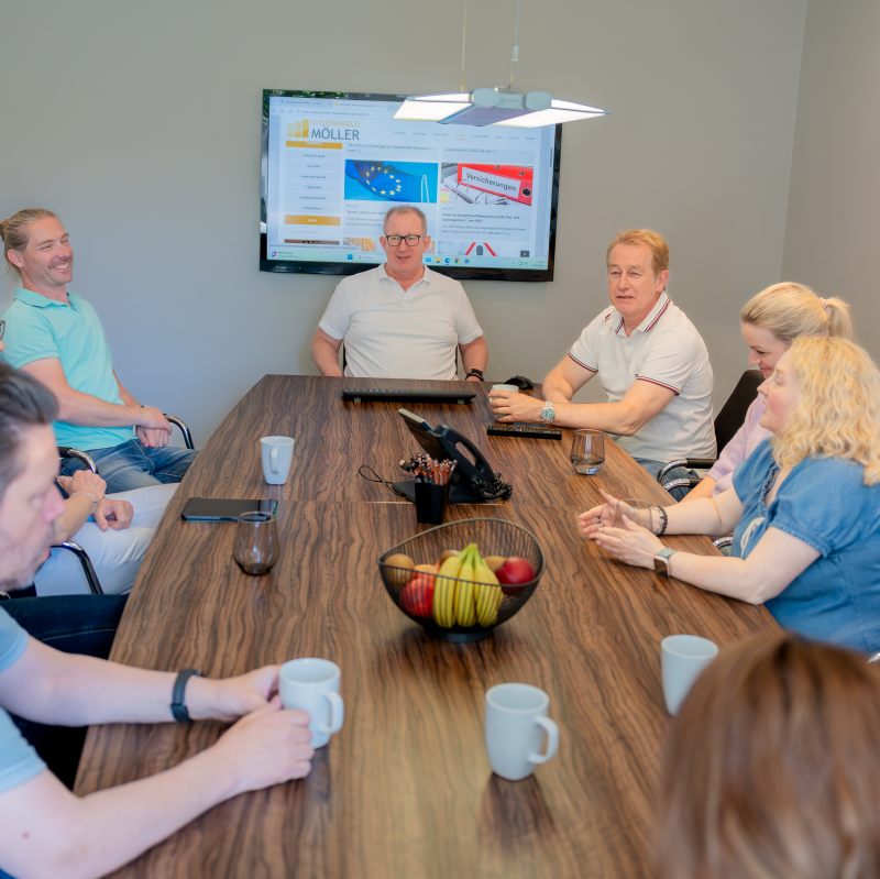 Sechs Personen sitzen um einen Konferenztisch, unterhalten sich, mit Tassen auf dem Tisch und einem Bildschirm im Hintergrund, der eine Präsentation zeigt.