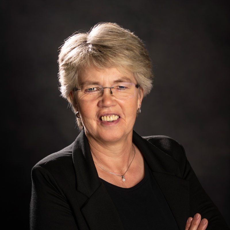 Steuerfachangestellte Maria Frye mit kurzen, grauen Haaren, Brille und schwarzem Blazer lächelt vor dunklem Hintergrund.