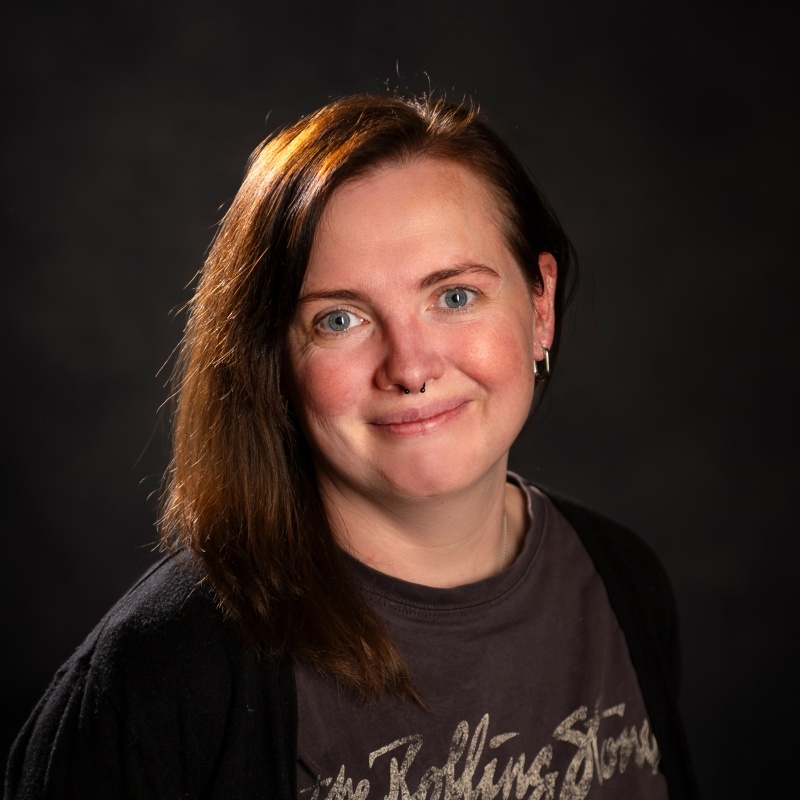 Steuerfachangestellte Tanja Schnieder mit schulterlangem braunem Haar und T-Shirt lächelt vor dunklem Hintergrund in die Kamera.